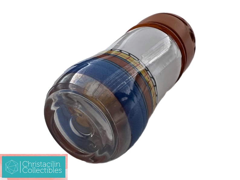 May include: A clear glass bottle with a brown cap. The bottle has a patterned design with blue, white, and yellow stripes. The bottle is cylindrical and appears to be a decorative item.