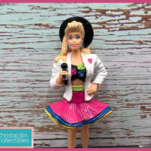 May include: A miniature figurine of a blonde woman wearing a white jacket, pink skirt, and black hat, holding a microphone. The figurine has blue eyes and is posed as if singing. The figure is a collectible item.