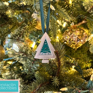 May include: A Christmas tree ornament shaped like a tree with a clock face. The clock face is green with white numbers and hands, and the brand name "Brookstone" is visible. The ornament hangs from a green ribbon and is set against a Christmas tree.