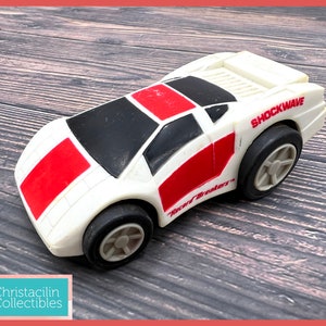 May include: A white and red toy car with black accents and the word "SHOCKWAVE" printed on the side. The toy car has black tires and the text "Record Breakers" is also visible. The car is a vintage collectible.