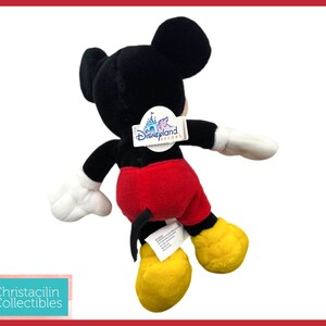 May include: A plush Mickey Mouse toy, viewed from the back, with black fur, red shorts, yellow shoes, and white gloves. The toy has a Disneyland Resort tag attached. The background is white with a red border.