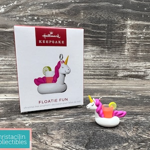 May include: A Hallmark Keepsake Christmas tree ornament featuring a white unicorn floatie with pink accents, a yellow horn, and a cocktail. The ornament is next to its box, which reads "FLOATIE FUN".