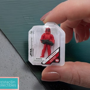 May include: A red Sith Trooper action figure from the Star Wars Toybox series, packaged in a clear plastic case. The figure is holding a black weapon. The packaging includes the text "Sith Trooper" and "15".