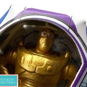Puede incluir: Figura de acci&oacute;n de Buzz Lightyear dorada en su embalaje original. La figura est&aacute; dentro de una burbuja de pl&aacute;stico transparente, con la caja con detalles en morado y azul. El embalaje incluye texto y el logotipo de Christacilin Collectibles.