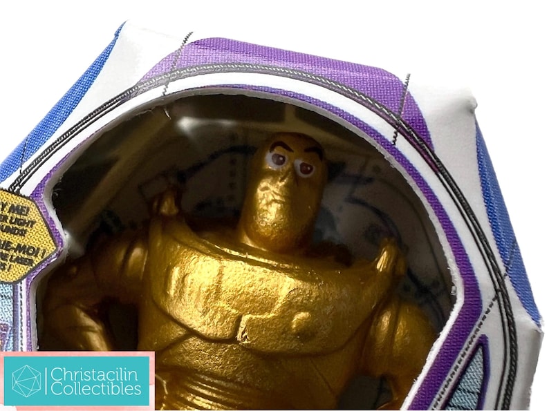 Puede incluir: Figura de acci&oacute;n de Buzz Lightyear dorada en su embalaje original. La figura est&aacute; dentro de una burbuja de pl&aacute;stico transparente, con la caja con detalles en morado y azul. El embalaje incluye texto y el logotipo de Christacilin Collectibles.
