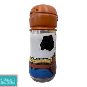 May include: A clear glass container with a brown lid, featuring a cartoon design. The container is decorated with a white background and a black cowhide pattern, with blue and brown accents. The design is reminiscent of a popular animated character.