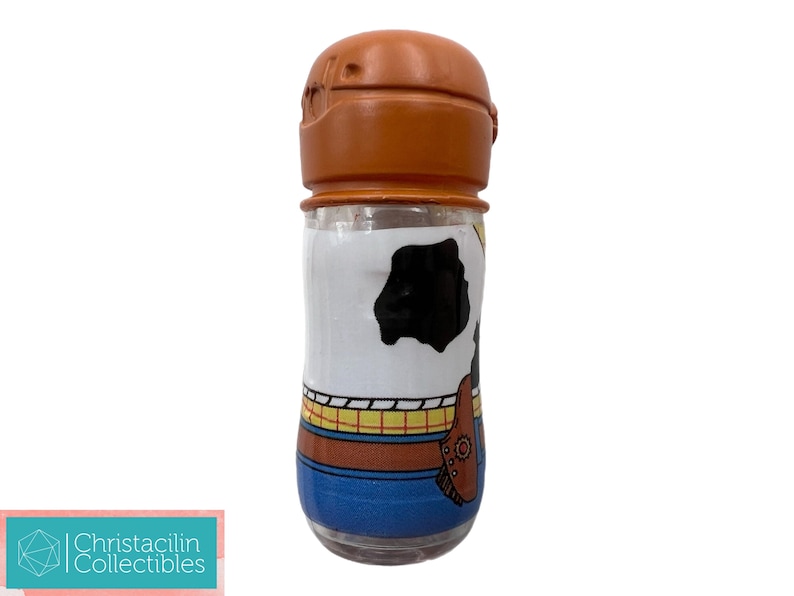 May include: A clear glass container with a brown lid, featuring a cartoon design. The container is decorated with a white background and a black cowhide pattern, with blue and brown accents. The design is reminiscent of a popular animated character.