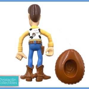 May include: A Woody action figure from the Toy Story franchise. The figure is wearing a cowboy hat, blue jeans, a yellow shirt, and brown boots. The figure is approximately 6 inches tall.