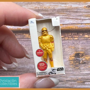 May include: A gold-colored Star Wars First Order Stormtrooper figurine in a white box. The box has the Star Wars logo and the Disney logo. The figurine is in a standing pose. The box has text that says "Blaster Lights and Sounds".