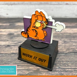 May include: A Garfield trophy figurine. The orange cat is depicted in a workout pose, with a white cloud emanating from his mouth. The trophy base is black with a gold plate that reads "WORK IT OUT".