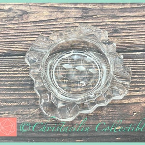 May include: A clear, vintage glass ashtray with a scalloped edge and diamond-shaped detailing in the center. The ashtray is circular and sits on a wooden surface. The text "Christacilin Collectibles" is visible.