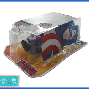 Op de afbeelding: Een Captain America actiefiguur in de originele verpakking. De figuur zit in een doorzichtige plastic hoes, met het Captain America schild zichtbaar. De verpakking heeft een rood-wit waarschuwingslabel. Van Christacilin Collectibles.