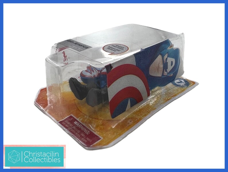 Op de afbeelding: Een Captain America actiefiguur in de originele verpakking. De figuur zit in een doorzichtige plastic hoes, met het Captain America schild zichtbaar. De verpakking heeft een rood-wit waarschuwingslabel. Van Christacilin Collectibles.