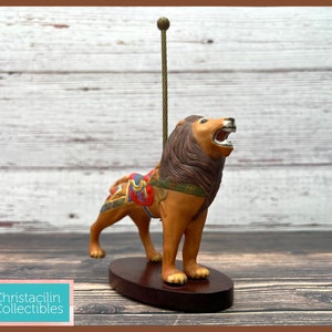 May include: A decorative lion figurine with a brown mane and open mouth, standing on a dark wooden base. The lion has a colorful saddle and a gold-colored pole extends upwards. The figurine is a collectible item.
