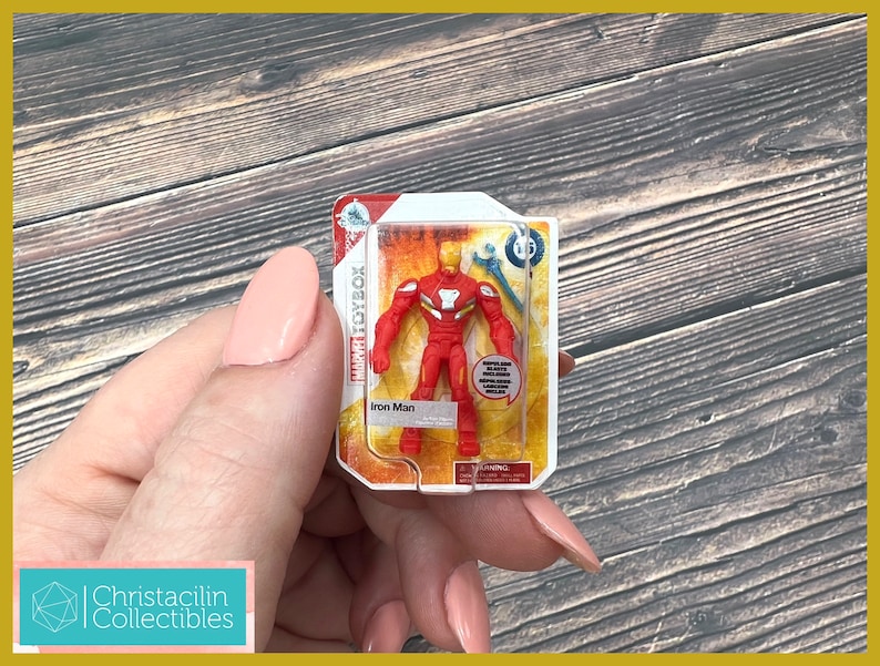 May include: A Marvel Toybox Iron Man action figure in a clear plastic package. The red and gold figure is visible, along with the packaging text "Iron Man". The package is held against a wooden background.