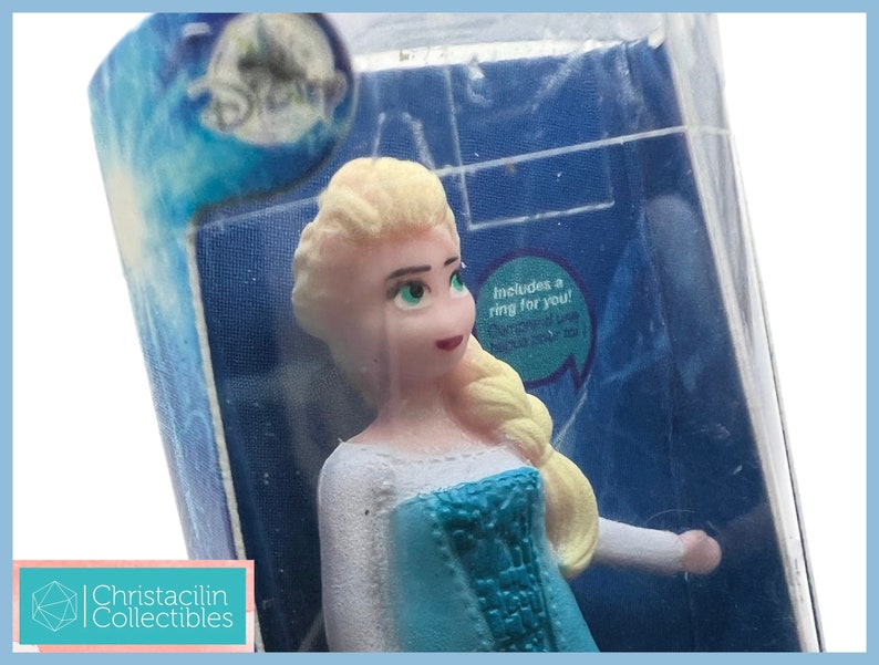 Pu&ograve; includere: Una piccola statuina di Elsa del film Disney Frozen, in una confezione di plastica trasparente. La statuina ha i capelli biondi, un vestito blu e occhi verdi. La confezione include il logo Disney e il testo "Includes a ring for you!"