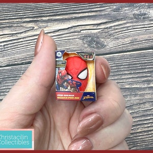 May include: A miniature Spider-Man mask in a box. The mask is red with white eye details. The box has the words "Spider-Man Mask" and "Masque de Spider-Man". The box is held in a hand.