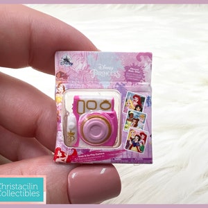 May include: A miniature pink and white Disney Princess Snap & Go Play Camera toy. The packaging features Disney Princesses and the Disney logo. The camera has a gold-colored lens and a pink body. The packaging includes images of Disney Princesses.