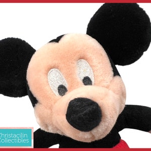 May include: Close-up of a Mickey Mouse plush toy. The plush features black ears, a black nose, and a cream-colored face. The toy has embroidered eyes and a red and black outfit. The image includes the text "Christacilin Collectibles".