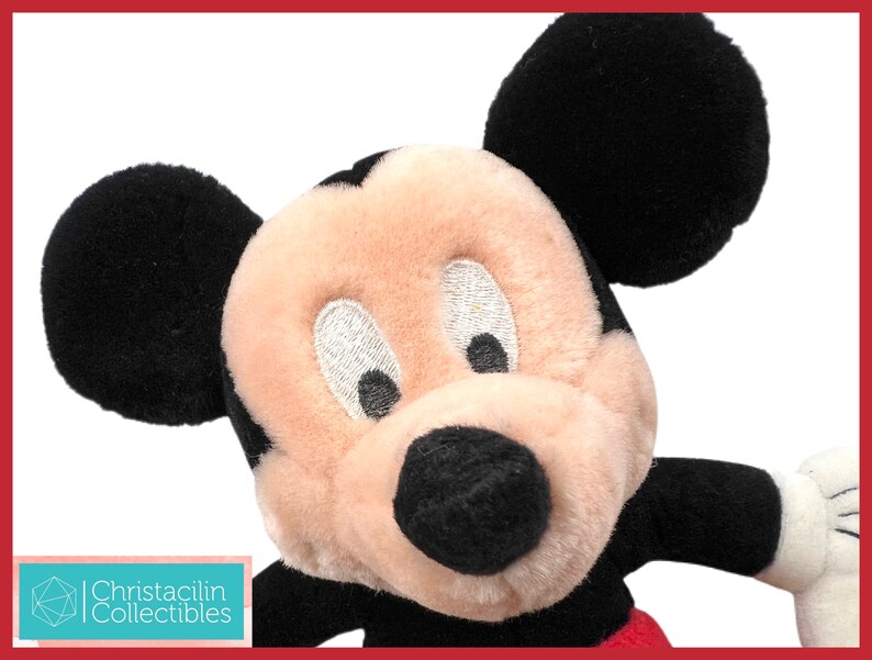 May include: Close-up of a Mickey Mouse plush toy. The plush features black ears, a black nose, and a cream-colored face. The toy has embroidered eyes and a red and black outfit. The image includes the text "Christacilin Collectibles".