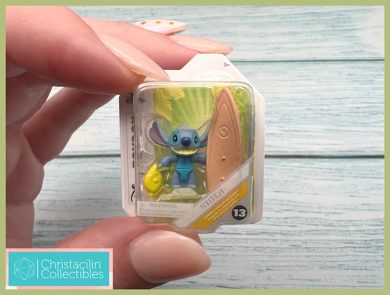 May include: A miniature Stitch figure from Disney's Lilo & Stitch, holding a surfboard and a yellow object, packaged in a clear plastic case. The figure is blue with large ears and a wide grin. The packaging includes the word "Stitch" and the number "13".