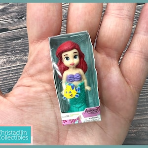 Op de afbeelding: Ariel-pop uit de Animators' Collection, in een doorzichtige plastic doos. De pop heeft rood haar, een paarse top en een groene staart. Er is ook een gele vis zichtbaar. De verpakking heeft de woorden "Animators' Collection" en "WARNING".