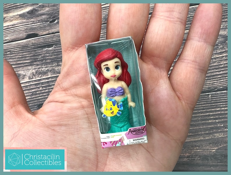 Op de afbeelding: Ariel-pop uit de Animators' Collection, in een doorzichtige plastic doos. De pop heeft rood haar, een paarse top en een groene staart. Er is ook een gele vis zichtbaar. De verpakking heeft de woorden "Animators' Collection" en "WARNING".
