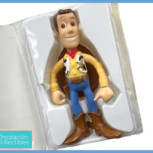May include: A Woody action figure from the Toy Story franchise, in its original packaging. The figure is wearing a yellow shirt, blue pants, a red bandana, and brown boots. The packaging is white and the figure is in a standing position.