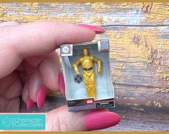 May include: A miniature gold-colored C-3PO figure from Star Wars in its original packaging. The figure is in a clear plastic case inside a small cardboard box. The box has the Star Wars logo and other text.