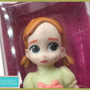 May include: A close-up of a figurine of a young girl with red hair in pigtails. She is wearing a light green dress with a pink bow. The figurine is in a clear plastic package with a purple background. The text "Christacilin Collectibles" is visible.