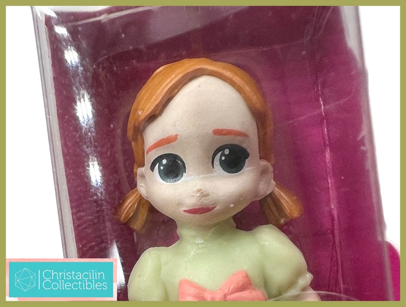 May include: A close-up of a figurine of a young girl with red hair in pigtails. She is wearing a light green dress with a pink bow. The figurine is in a clear plastic package with a purple background. The text "Christacilin Collectibles" is visible.