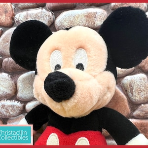 May include: A plush Mickey Mouse doll with black ears, a black nose, and a red and white outfit. The doll has a cream-colored face and is set against a stone background. The doll is a collectible item.