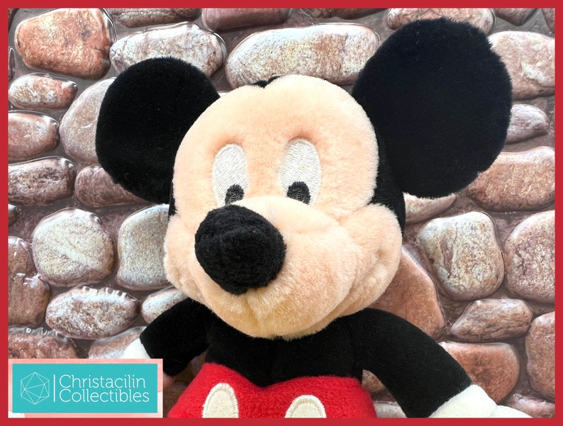 May include: A plush Mickey Mouse doll with black ears, a black nose, and a red and white outfit. The doll has a cream-colored face and is set against a stone background. The doll is a collectible item.