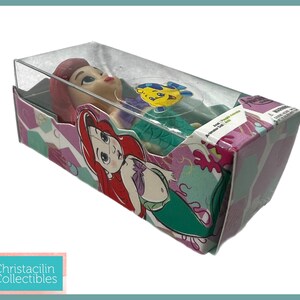 Op de afbeelding: Ariel-pop in een doorzichtige plastic doos. De doos heeft een kleurrijk ontwerp met afbeeldingen van Ariel en Botje. De pop heeft rood haar en een groene staart. De doos is gelabeld met "Ariel, De Kleine Zeemeermin".