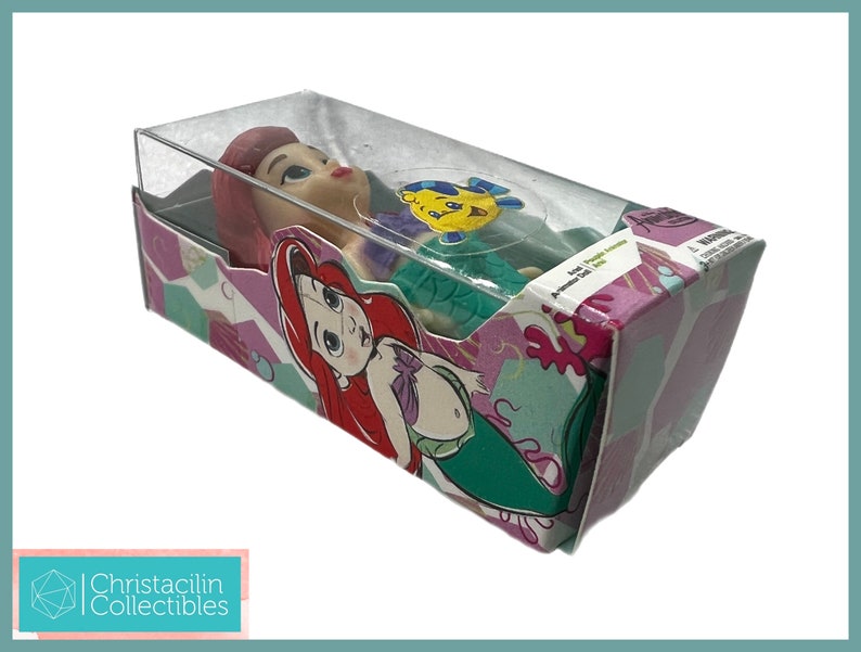 Op de afbeelding: Ariel-pop in een doorzichtige plastic doos. De doos heeft een kleurrijk ontwerp met afbeeldingen van Ariel en Botje. De pop heeft rood haar en een groene staart. De doos is gelabeld met "Ariel, De Kleine Zeemeermin".