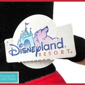 May include: A black plush toy with a red and white design, holding a Disneyland Resort tag. The tag features a blue castle illustration and the Disneyland Resort logo. The toy's design is reminiscent of a classic cartoon character.