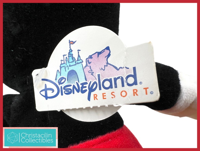 May include: A black plush toy with a red and white design, holding a Disneyland Resort tag. The tag features a blue castle illustration and the Disneyland Resort logo. The toy's design is reminiscent of a classic cartoon character.