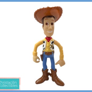 May include: A Woody action figure from the Toy Story franchise. The figure is wearing a brown cowboy hat, a yellow shirt, a red bandana, blue jeans, and brown boots. The figure has a sheriff's badge and a holster.