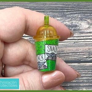 May include: A miniature green and yellow plastic cup with a lid, featuring the text "SNACK ATTACK" and a cartoon character. The cup has a green patterned design and is held in a person's hand.