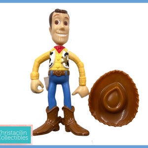 May include: A Woody action figure from the Toy Story franchise, with a brown cowboy hat. The figure is wearing a yellow shirt, blue pants, and brown boots. The figure has a sheriff's badge and a red bandana.