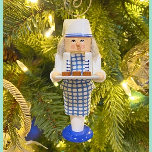 May include: A wooden Christmas ornament of a chef holding a tray of pastries. The chef is wearing a white hat and uniform with blue accents. The ornament has a gold wire for hanging and is set against a Christmas tree background.