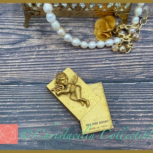 May include: A gold-colored matchbox with a relief of a cherub playing a harp. The matchbox is open, revealing the matches inside. The text "GOLD WOOD MATCHES MADE IN ITALY" is visible. A pearl necklace and a small gold-colored jewelry box are in the background.