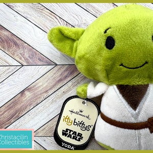 May include: A green plush Yoda doll from Star Wars, with large ears and a smiling face. The doll wears a white robe with a brown belt. The tag reads "itty bittys" and "Star Wars Yoda".