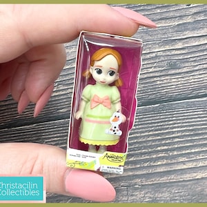 May include: A miniature doll of a character from the movie Frozen, packaged in a clear plastic box. The doll has a light green dress with a pink bow and a small Olaf figure. The box has the words "Animators Collection" printed on it.