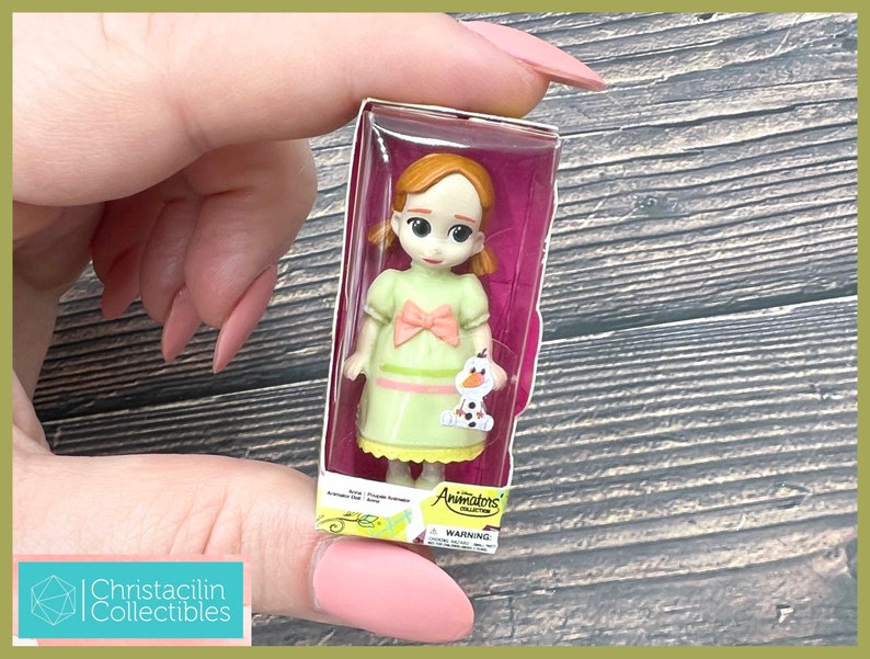 May include: A miniature doll of a character from the movie Frozen, packaged in a clear plastic box. The doll has a light green dress with a pink bow and a small Olaf figure. The box has the words "Animators Collection" printed on it.