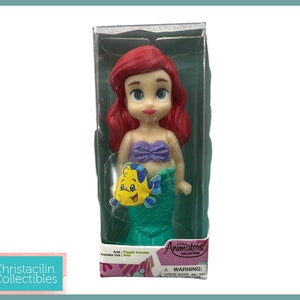 Op de afbeelding: Ariel pop uit de Animators' Collection in een doorzichtige plastic doos. De pop heeft rood haar, een paarse top en een groene staart. Een gele vis is aan de staart bevestigd. Op de doos staan de woorden "Animators' Collection" en "Ariel".