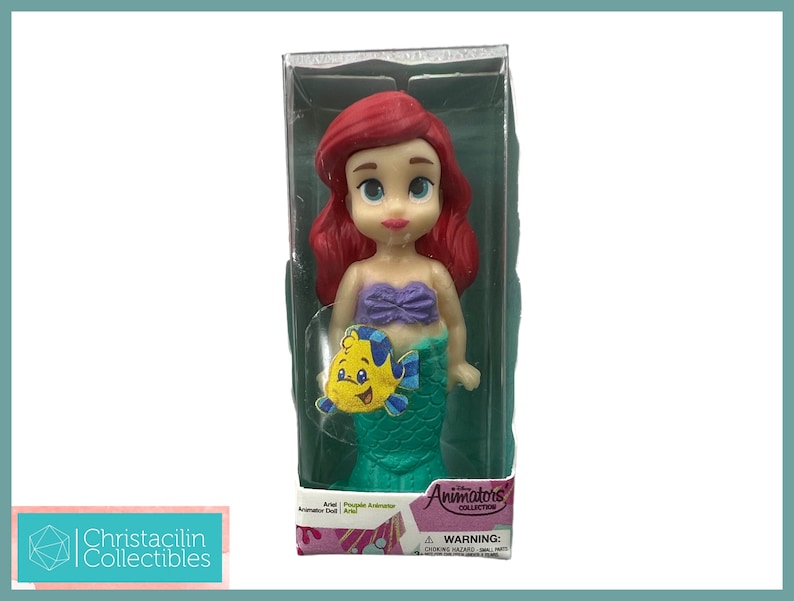 Op de afbeelding: Ariel pop uit de Animators' Collection in een doorzichtige plastic doos. De pop heeft rood haar, een paarse top en een groene staart. Een gele vis is aan de staart bevestigd. Op de doos staan de woorden "Animators' Collection" en "Ariel".