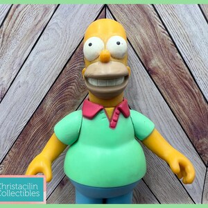 May include: A Homer Simpson figurine with a yellow head, a light green shirt, and blue pants. The figure has a large smile and a red collar. The background is a wooden chevron pattern.