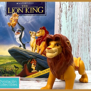 May include: A Lion King Simba figurine in front of a VHS tape of the movie. The VHS tape features the movie title and characters. The Simba figurine is yellow and brown. The background is a light blue and white wooden surface.