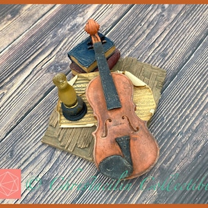 May include: A detailed figurine featuring a violin, sheet music, and small books. The violin is brown with a black fingerboard, and the books are blue and brown. The figurine is set on a wooden surface.